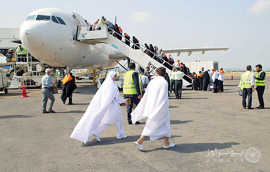 مسافران حج درحال سوار هواپیما شدن در فرودگاه ایران؛ دو مسافر سفیدپوش زن و مرد در جلوی تصویر و چند نفر از مسئولان فرودگاه با جلیقه شب‌رنگ دیده می‌شوند.